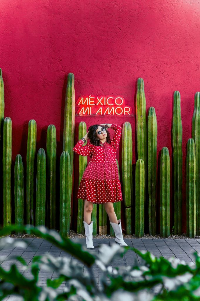 woman wearing dress and sunglasses standing in front of cactus and pink wall