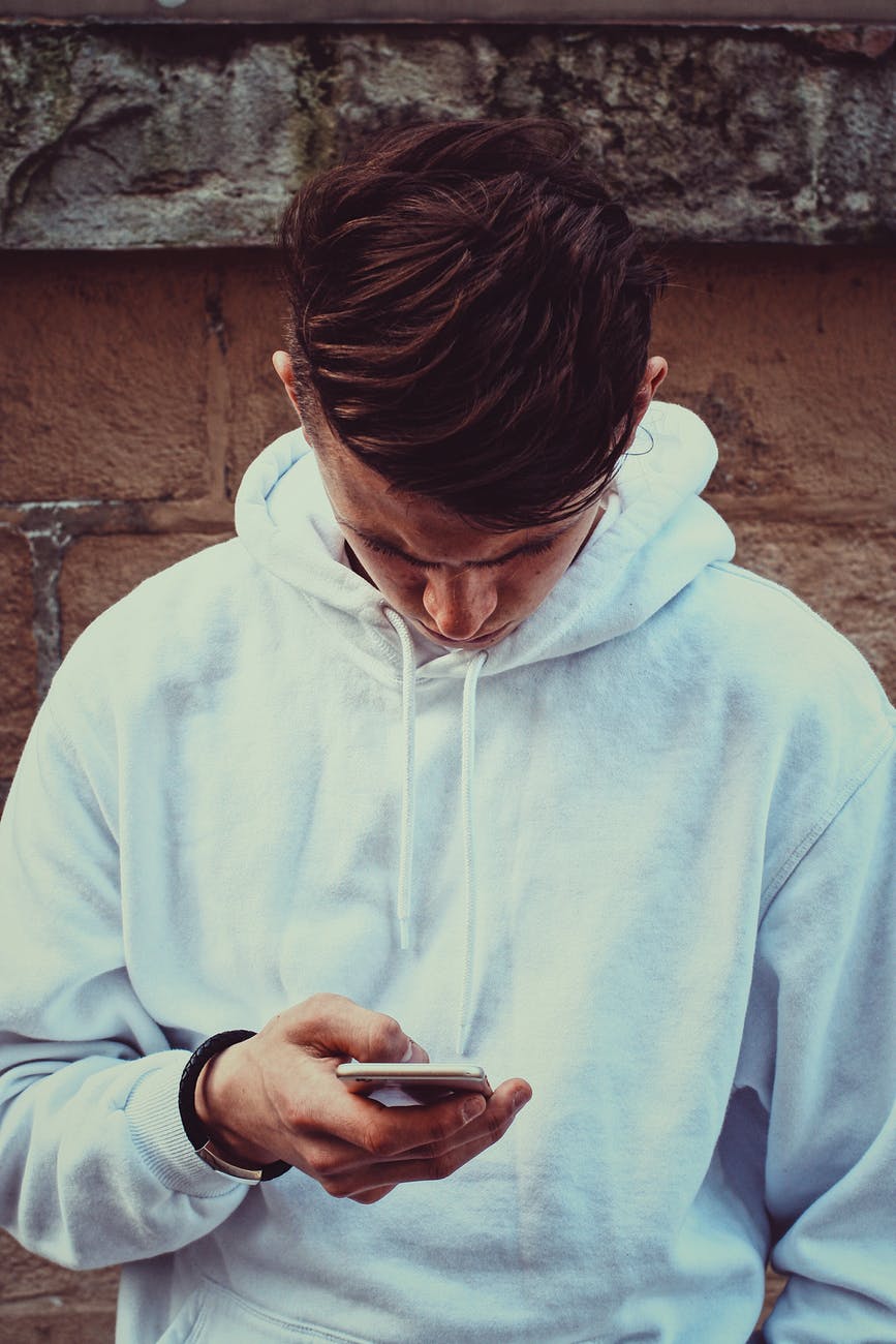 photo of man in white hoodie standing near brown wall while using his smartphone